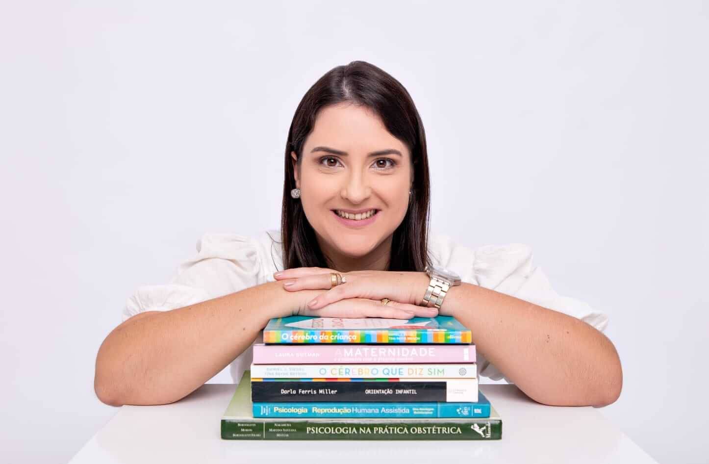 Dany Monteiro sorrindo, com os braços apoiados sobre uma pilha de livros de psicologia